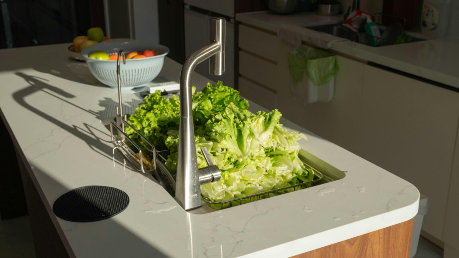 A sunlit modern kitchen counter with fresh lettuce and fruit bowl near a shiny faucet.